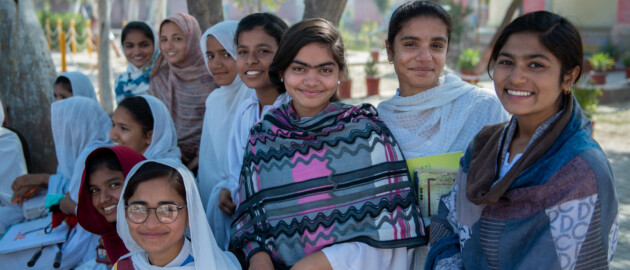 Adolescent girls stand outside in a group and smile and laugh looking at the camera.