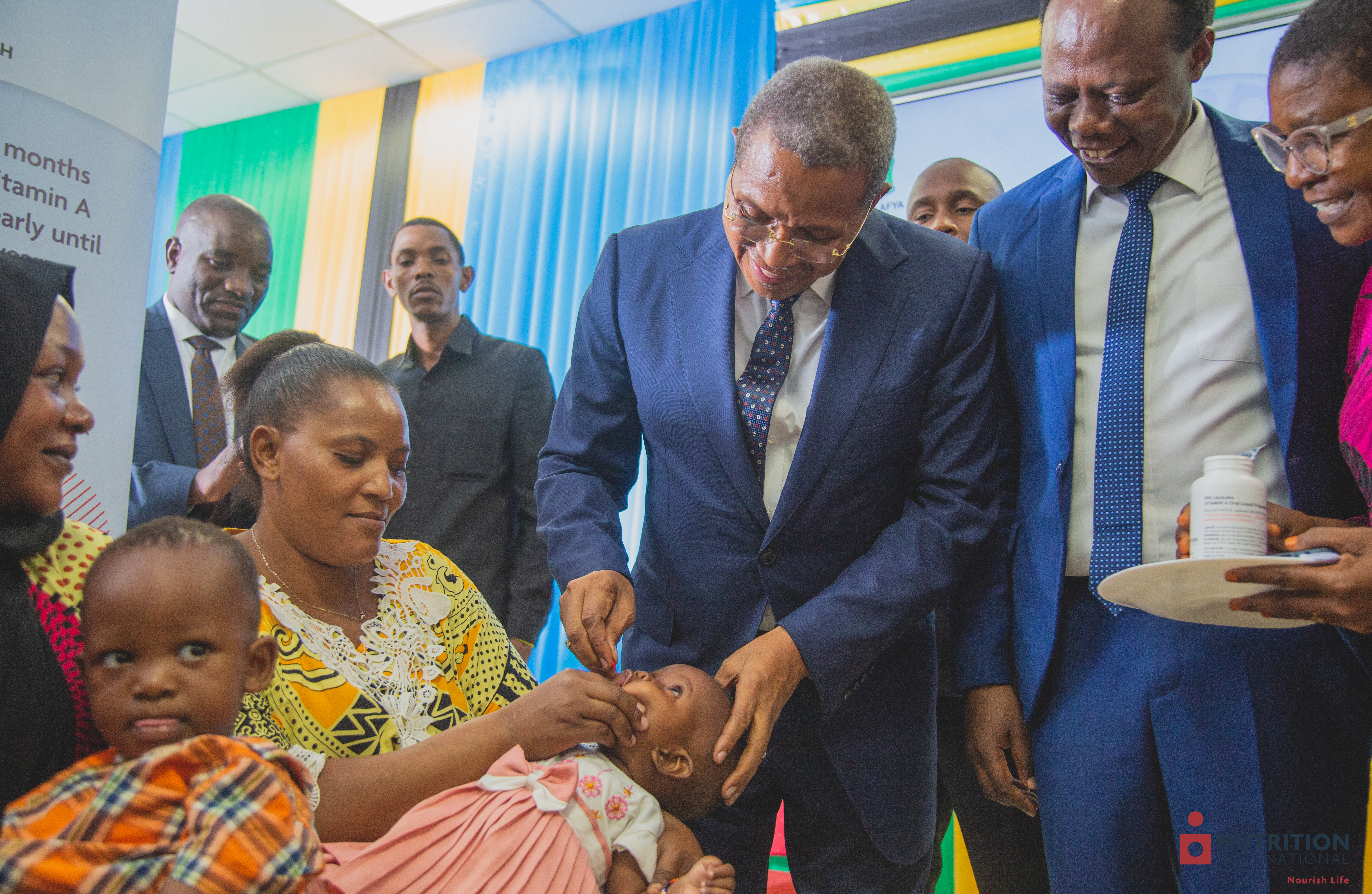 A man administers a red supplement to a child in its mother's arms.