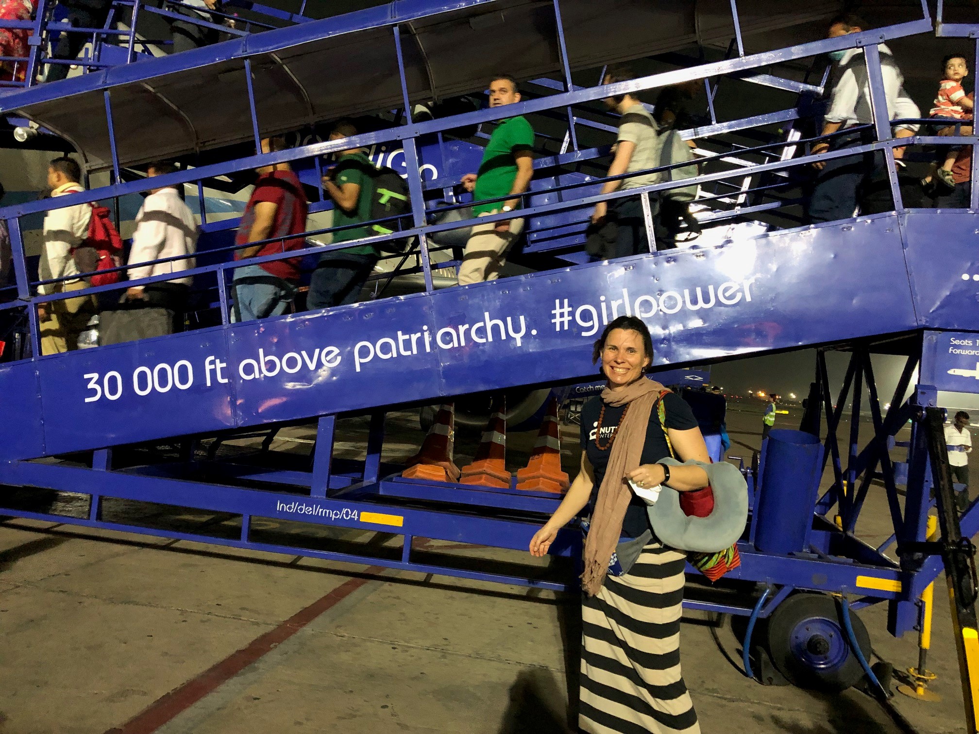 A woman stands outside on the tarmac of a airport. People are boarding a plane through an outdoor staircase.