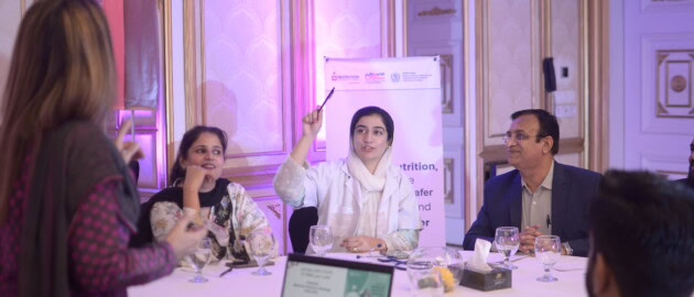 People discuss across a table in a room. A woman is raising her hand to add to the discussion.