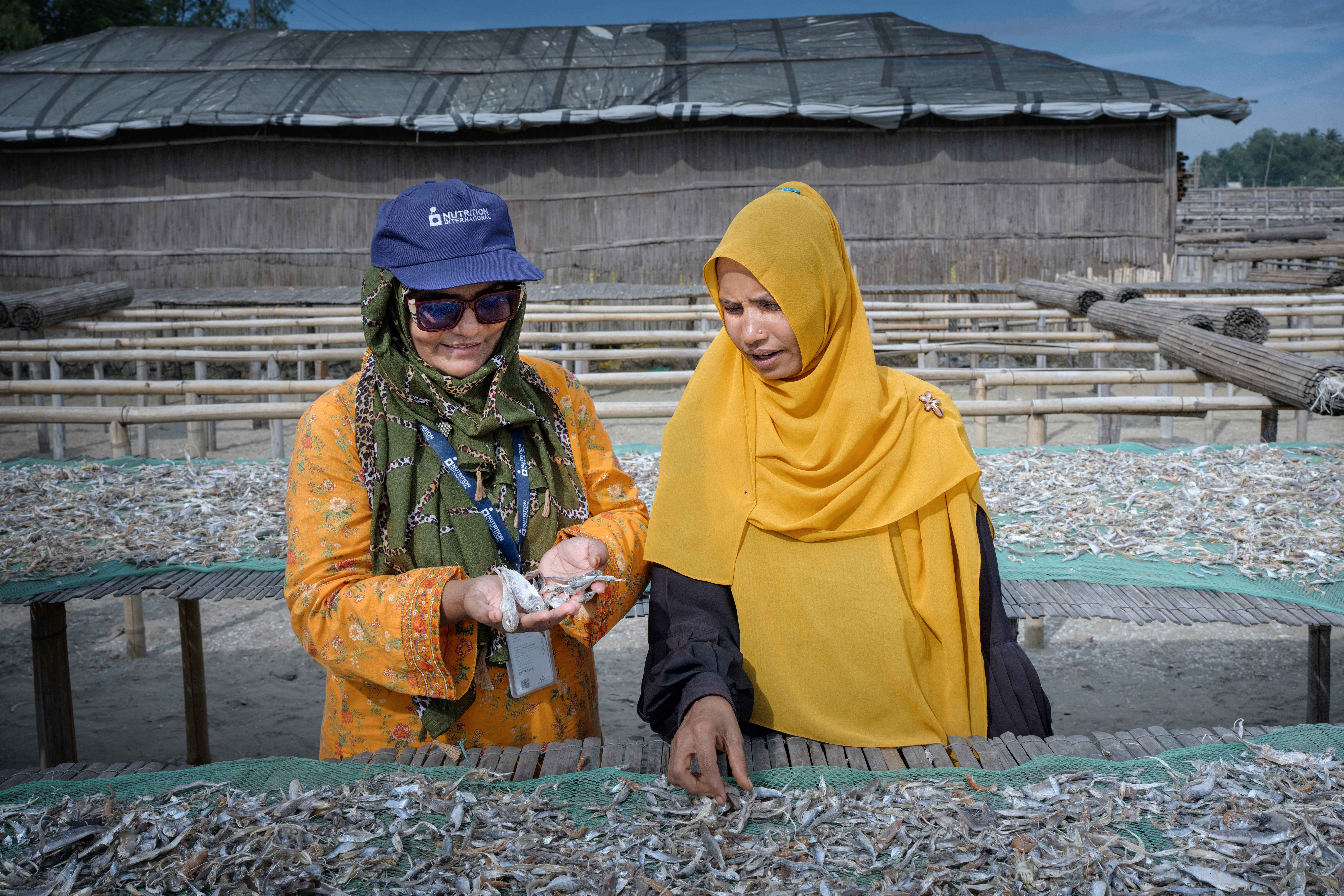 Two woman stand in front of a row of fish. One woman is explaining what's happening to the other woman.