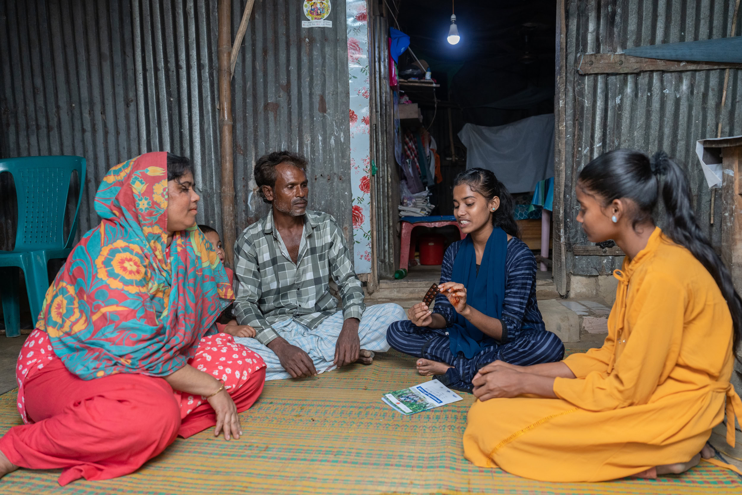 A family sits in a semi circle. An adolescent girl holds up a package of weekly iron and folic acid supplementation and is speaking to her family.